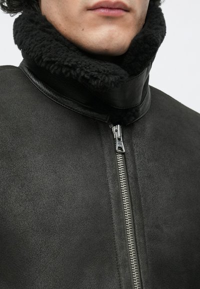 Juoda rauginta avių kailio striukė su aukštu apykakliu, užtrauktuku ir minkšta, tekstūruota medžiaga. Detalėse matoma lygi odinė vidinė apykaklė.