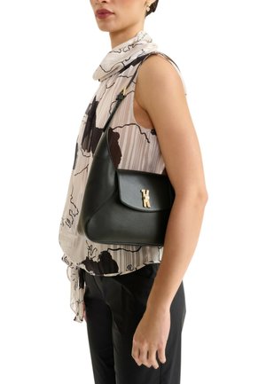 Donna che indossa una blusa smanicata con fantasia e pantaloni neri, che porta una borsa a tracolla in pelle nera con una chiusura a logo dorato.