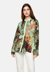Brown shirt featuring a green leafy print, long sleeves, a collar, and a green checkered button placket. White pants complete the outfit.