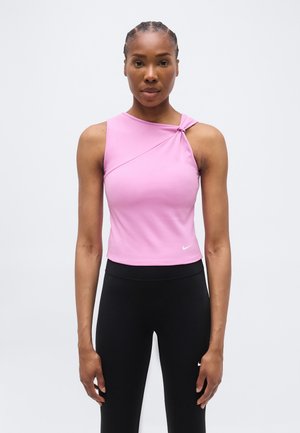 Žena čelom dopredu, oblečená v ružovom tielku bez rukávov s uzlom na jednom ramene a čiernych legínach s vysokým pásom.