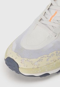 Scarpa da ginnastica bianca con tomaia in mesh, dettagli testurizzati e intersuola giallastra con design macchiato. Suola in gomma scura visibile.