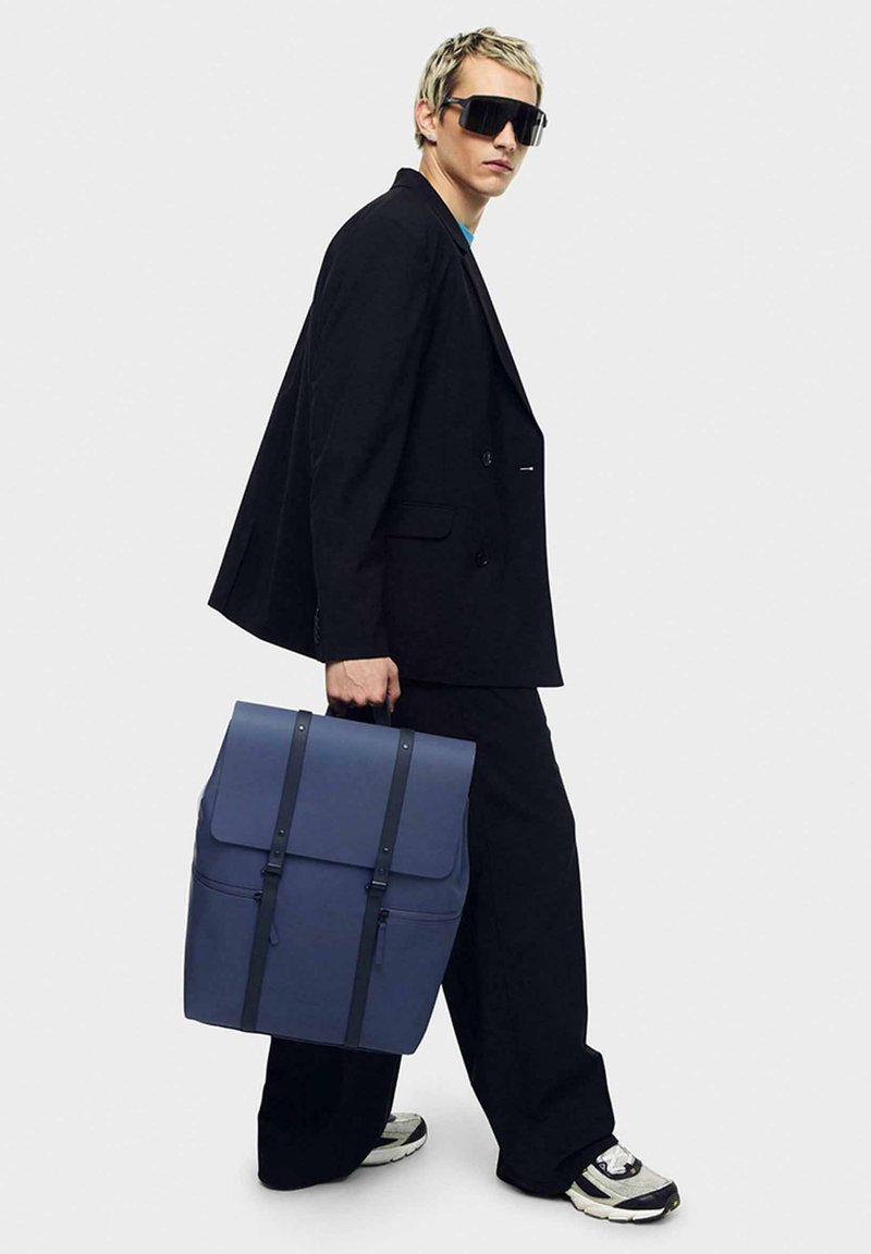 Marinblå ryggsäck med klaff, dubbla axelremmar och framficka med dragkedja. Slät material, strukturerad form och minimalistisk design.