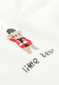 Tela blanca con un oso bordado que lleva un abrigo rojo y un sombrero negro. Debajo, está bordado en negro "pequeño oso".