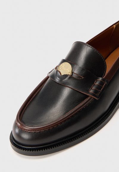 Svart läderloafer med en brun rem och guld Clarks-logga. Har en sydd söm och en texturerad svart sula. Rundad tå.