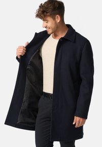 Mørkeblå uldfrakke med skjortekrave, knaplukning og en glat sort indvendig foring. Modellen har en lys beige sweater på under.