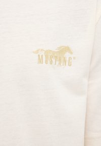 Tissu beige clair avec un petit logo doré représentant un cheval au galop et le mot "MUSTANG" sur la partie supérieure de la poitrine.