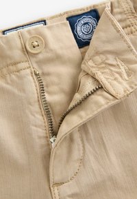 Pantaloni di cotone beige con chiusura a zip in ottone e bottone. Presentano un'etichetta in tessuto con sfondo blu e testo, e cuciture pulite sui bordi.