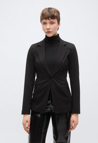 Μαύρο ραμμένο σακάκι με ένα κουμπί, γιακάδες με σχισμή και πλαϊνές τσέπες, συνδυασμένο με μαύρο ζιβάγκο και γυαλιστερό μαύρο παντελόνι.