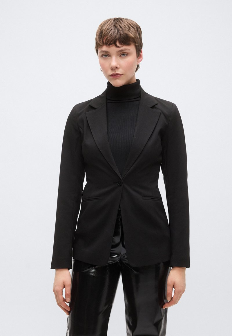 Μαύρο ραμμένο σακάκι με ένα κουμπί, γιακάδες με σχισμή και πλαϊνές τσέπες, συνδυασμένο με μαύρο ζιβάγκο και γυαλιστερό μαύρο παντελόνι.