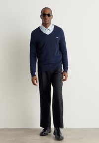 Pull en V marin sur une chemise à col bleu clair, associé à un pantalon noir et des chaussures noires, agrémenté de lunettes de soleil noires.