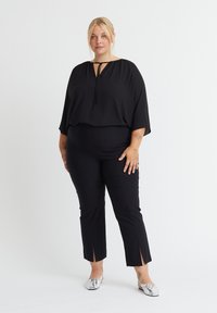 Schwarze, locker sitzende Bluse mit rundem Ausschnitt und Bindedetail, kombiniert mit schwarzen, schmalen Hosen mit Seitenschlitzen und silbernem Schuhwerk.