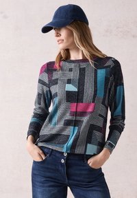 Barevný svetr s dlouhým rukávem se geometrickými vzory v černé, tyrkysové a růžové barvě. Materiál vypadá strukturovaně. Spárováno s tmavě modrými džíny.