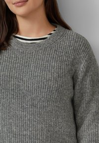 Grijze gebreide sweater met een gemêleerde afwerking, voorzien van een ronde halslijn en zichtbare ribbels; gedragen over een zwart-wit gestreept shirt.