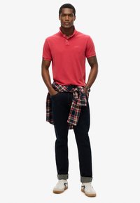 Polo rosso con colletto, abbinato a jeans scuri arrotolati alle caviglie e una camicia flanella a quadri legata in vita. Sneakers bianche.