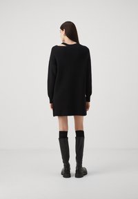 Abito maglia nera con maniche lunghe, vestibilità ampia e scollatura bassa sul retro. Indossato con stivali in pelle neri fino al ginocchio.