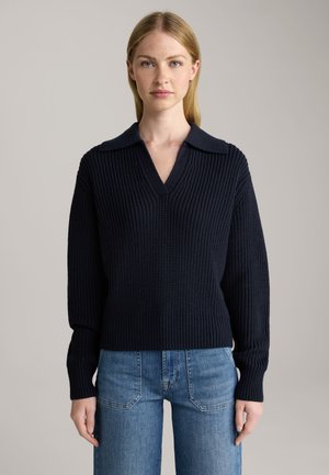 Pull en tricot bleu marine avec un col en V et un design à col. Il présente une texture côtelée et des manches longues, associé à un jean en denim bleu clair.