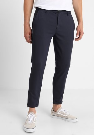 Uomo che indossa pantaloni slim fit blu scuro con cerniere alle caviglie, sneakers bianche e t-shirt bianca infilata, in piedi davanti a uno sfondo semplice.
