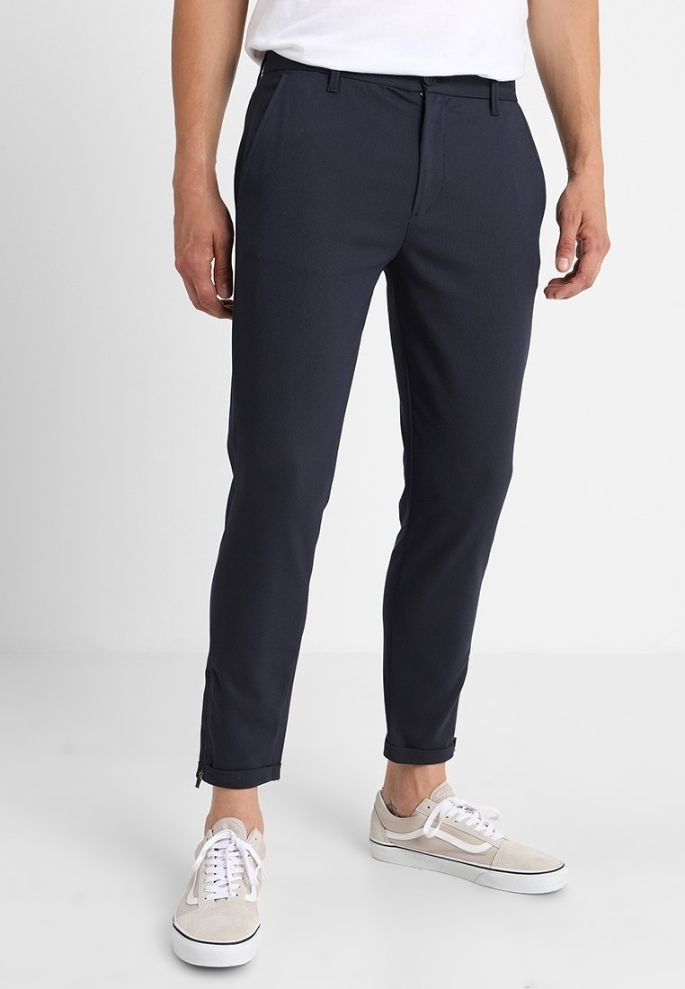 Homme portant un pantalon slim bleu marine foncé avec des fermetures éclair aux chevilles, des baskets blanches et un t-shirt blanc rentré, debout devant un fond uni.