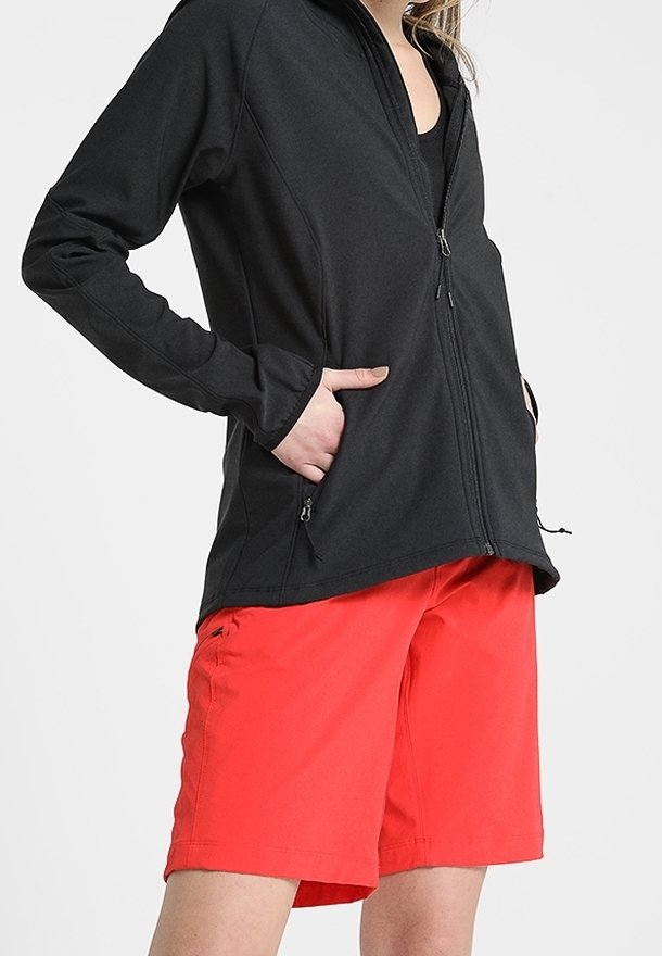 Personne portant une veste noire à fermeture éclair, les mains dans les poches, et un short rouge vif jusqu'aux genoux sur un fond uni.