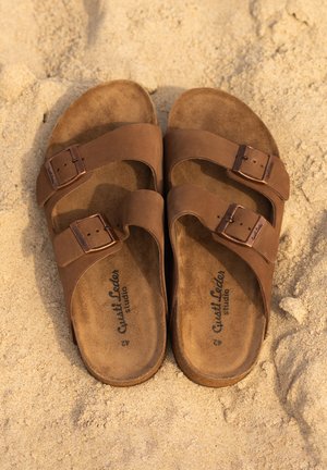 Bruine leren sandalen met twee verstelbare banden, metalen gespen en een zachte nubuckachtige binnenzool, rustend op zand. Bevat merklettertype.