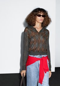 Grijze gebreide polo met argylepatroon en lange mouwen, gecombineerd met lichtblauwe denimjeans en een rode geknoopte trui om de taille.