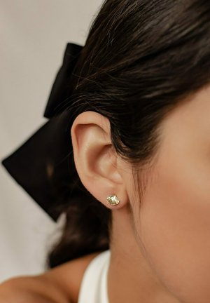 Goldblatt-förmiger Steckerohrring am durchstochenen Ohr. Das Design zeichnet sich durch strukturiertes Detail und eine polierte Oberfläche aus, die dunkelhaarige Trägerinnen ergänzt.