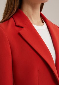 Červený blejzr s strukturovaným límcem, hladkou texturou tkaniny a slim fit střihem, k němuž je vrstvený bílý žebrovaný top.