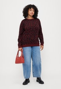 Sweater de malha com padrão de leopardo vermelho e preto, gola redonda, punhos e bainha canelados; combinado com jeans de perna larga azul claro e uma bolsa vermelha.