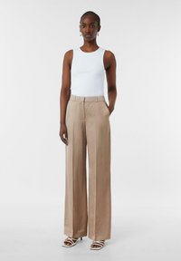 Beżowe spodnie o szerokich nogawkach w dopasowanym kroju, zestawione z białym, prążkowanym topem na ramiączkach. Modelka w białych sandałach na obcasie ze strunami.