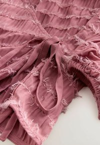 Rynket pink stof top med strukturerede striber, frynsede kanter og en bindedetalje. Designet har sammensamlet elastiske accenter i taljen.