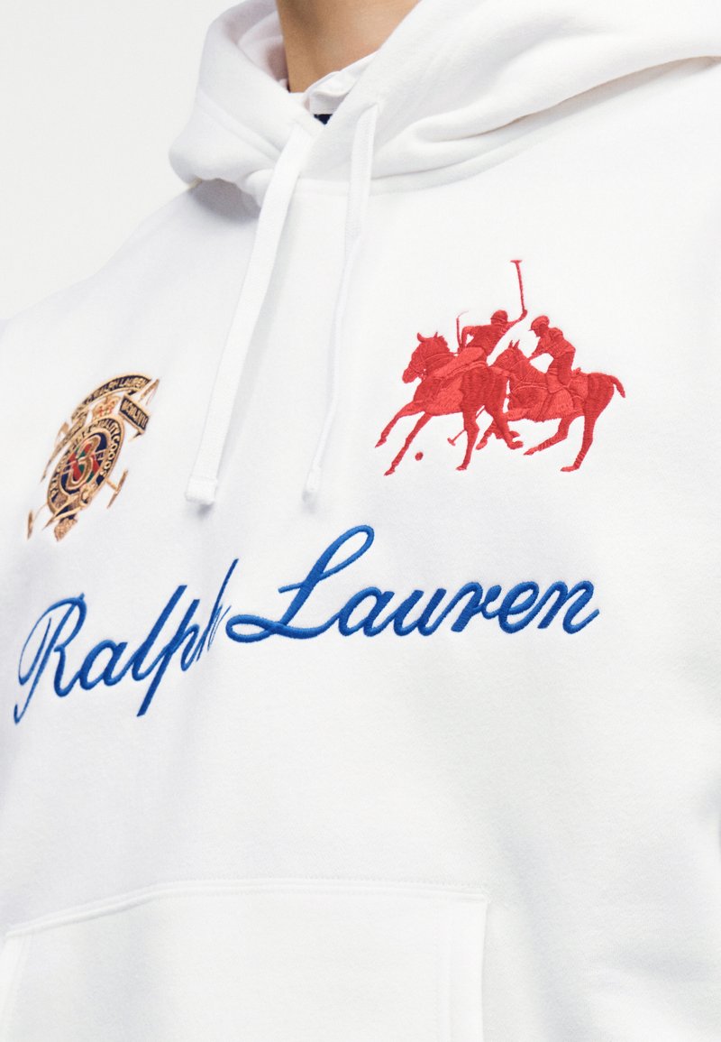 Sweat à capuche blanc avec l'inscription "Ralph Lauren" en bleu, joueurs de polo brodés en rouge à cheval, et un emblème brodé en or sur la poitrine.