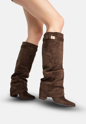 Benedenbenen met hoge, nonchalante donkerbruine suède laarzen met lage hakken en puntige neuzen, met een gouden gespdetail vlak bij de bovenkant.