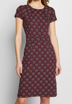 Vrouw draagt een knielange jurk met korte mouwen en een rood-zwart geometrisch bloemmotief, staand tegen een neutrale achtergrond.