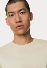 Lys beige T-shirt med rund halsudskæring, glat tekstur og korte ærmer. Stoffet virker blødt og tætsiddende omkring halsen.