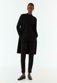 Schwarzer Cardigan, eng anliegendes schwarzes Top, maßgeschneiderte schwarze Hose und Slipper mit goldenen Beschlägen. Glatte Texturen und schlanke Silhouette durchgehend.