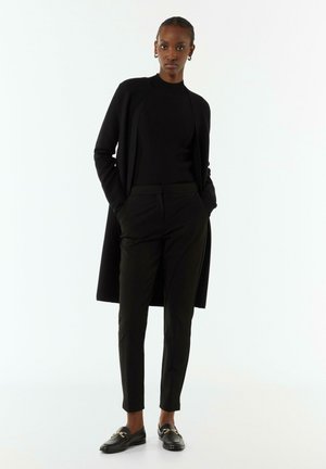 Zwarte cardigan, aansluitend zwart topje, op maat gemaakte zwarte broek en loafers met gouden accenten. Gladde texturen en slanke silhouetten door het geheel.
