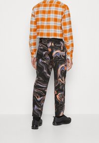 Man som bär svarta byxor med orange och lila virvlar, en orange rutig skjorta och svarta sneakers, som står mot en vit bakgrund.