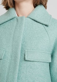 Cappotto in misto lana verde menta con superficie texturizzata, dotato di colletto appuntito e due tasche a toppa sul davanti.