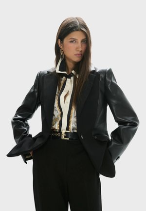 Mujer con pelo largo y castaño lleva una chaqueta negra con mangas de cuero, blusa estampada con cadenas doradas, pantalones negros de cintura alta y pendientes de aro dorados.
