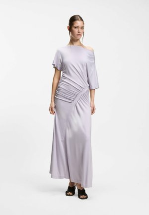 Kvinde iført en lyse lavendel, off-shoulder, rynket maxikjole og sorte åbne hælede sandaler stående mod en ensfarvet hvid baggrund.