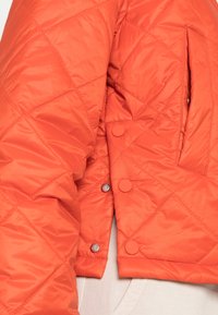 Giacca arancione trapuntata con chiusura a bottone a pressione, tasca laterale e texture liscia e lucida. Il design è accorciato sopra la vita.