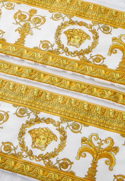 Tissu blanc avec des motifs dorés complexes, incluant des feuilles de laurier et un motif central de visage, bordé d'éléments décoratifs dorés.
