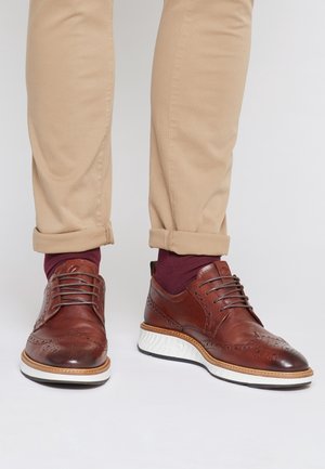 Brązowe brogsy z skóry z białą podeszwą noszone z beżowymi podwiniętymi spodniami i ciemnoczerwonymi skarpetkami na zwykłym jasnym tle.