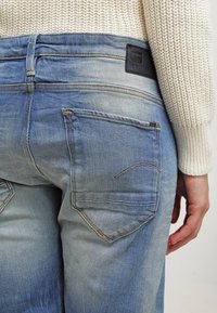 Ljusblåa jeans i denim med blekt tvätt, två bakfickor och en texturerad svart läderlogotyp på midjebandet. Har synliga sömmar.