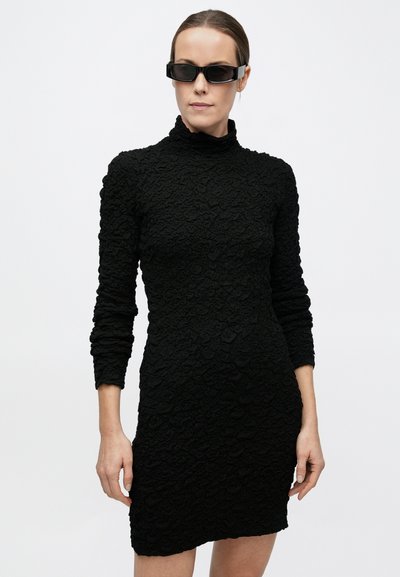 Naine, kes kannab tekstuuriga musta pika varrukaga minikleit ja musta ristküliku kujulisi päikeseprille, seisab ühevärvilise heleda tausta ees.