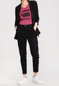 Sorte skræddersyede bukser, pink V-hals t-shirt med sort logo og hvide sneakers. En blazer med opsmøgede ærmer komplementerer outfittet.