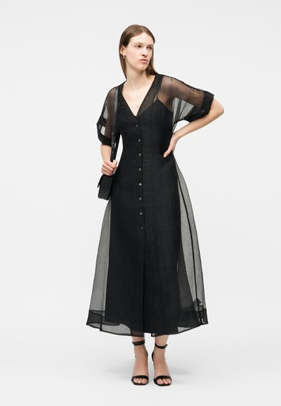 Γυναίκα που φοράει μαύρο midi φόρεμα με κουμπιά μπροστά, διαφανή κοντά μανίκια και επικάλυψη, κρατώντας μαύρη τσάντα ώμου και φορώντας μαύρα πέδιλα με λουριά.