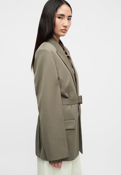 Μπλέιζερ σε ελαιόχρωμο πράσινο με στιβαρούς ώμους, φαρδιά μανίκια και ζώνη στη μέση. Διαθέτει κλείσιμο με ένα κουμπί και μπροστινές τσέπες. Απαλό ύφασμα.