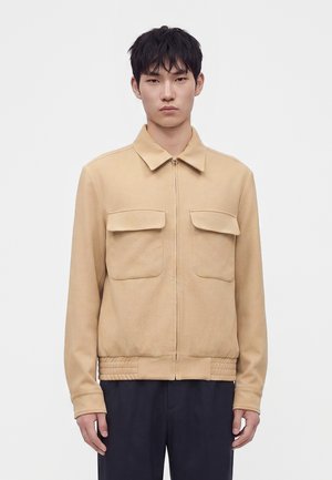Jeune homme portant une veste beige zippée avec deux poches à rabat à l’avant et un pantalon foncé, debout devant un fond blanc uni.