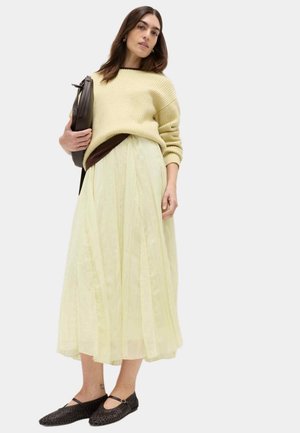 Donna con gonna midi giallo pallido, maglione giallo chiaro, ballerine nere intrecciate e che porta una borsa a tracolla scura, in piedi davanti a uno sfondo bianco.